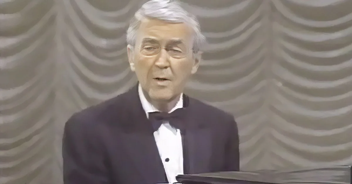 A man in a tuxedo and bow tie stands at a podium, speaking in front of a wavy-patterned backdrop.