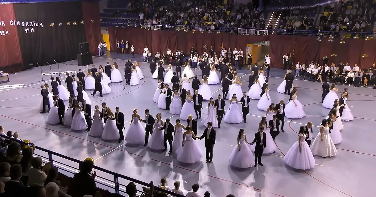 A grand ballroom scene with couples in formal attire dancing in a large circle.