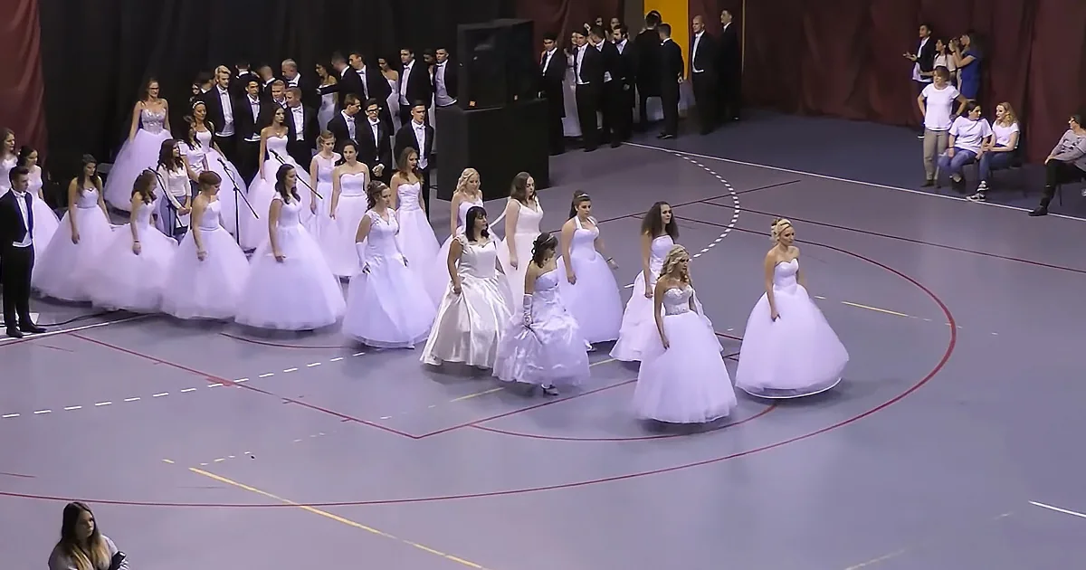 Young women in white dresses walk in a line on a basketball court.