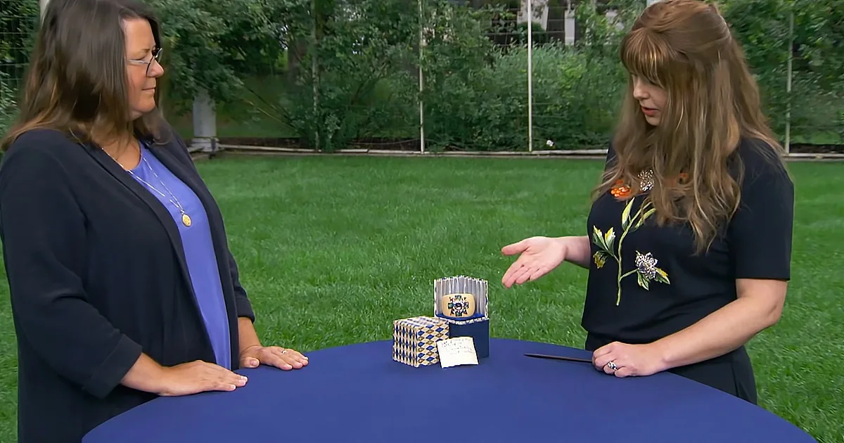 Two women stand at a table outdoors, one gesturing, with a stack of cards and a pen on the table.