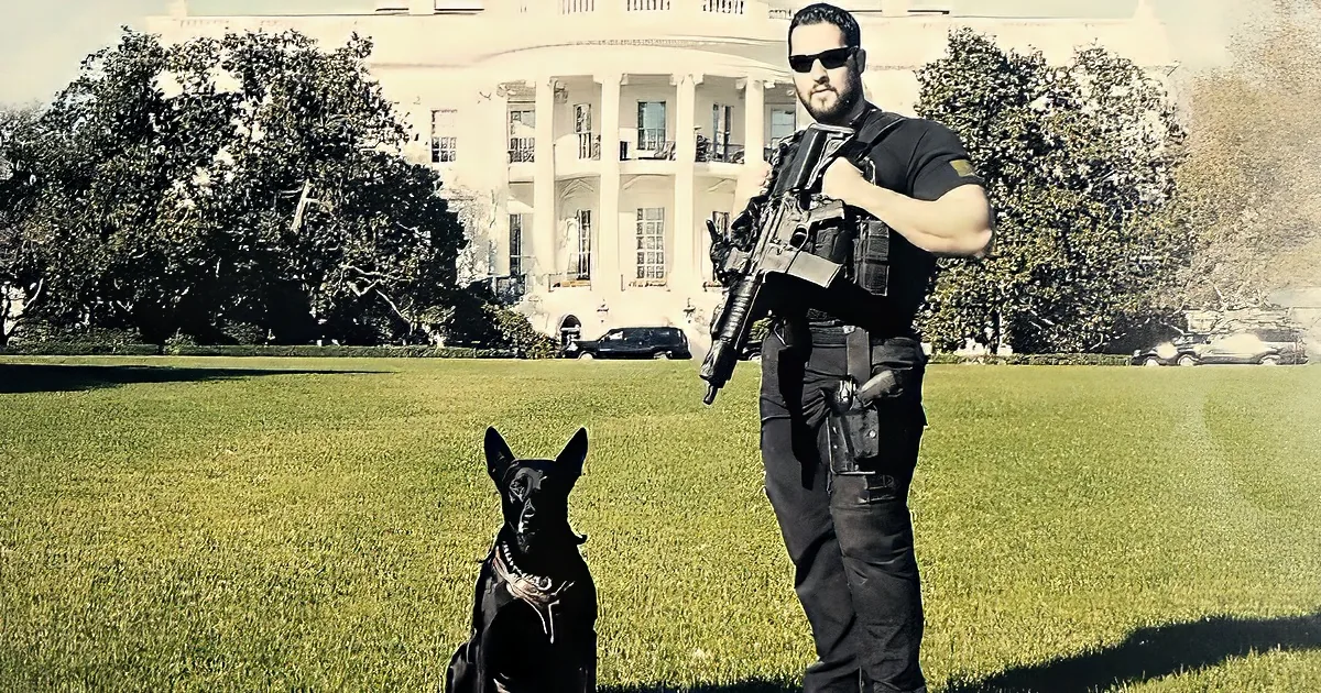 A man in tactical gear stands with a dog on the lawn in front of the White House.