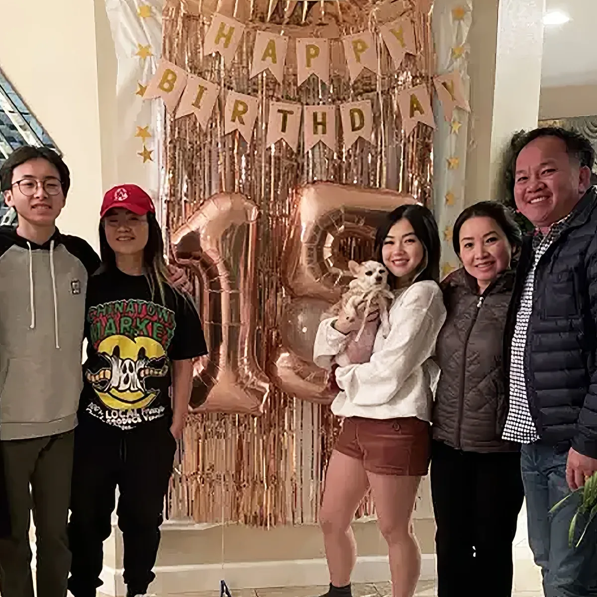 A group of five people, including a woman holding a dog, stands in front of a "Happy Birthday" banner and large number 20...