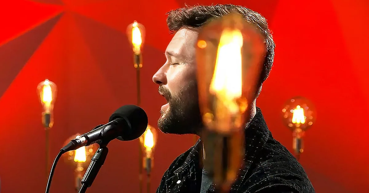 A man sings into a microphone, surrounded by glowing light bulbs against a red backdrop.