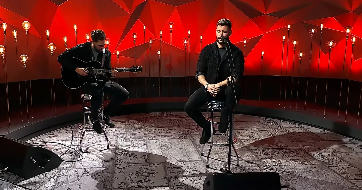 Two musicians perform on stools, one playing guitar, the other singing into a microphone, against a red geometric backdrop.