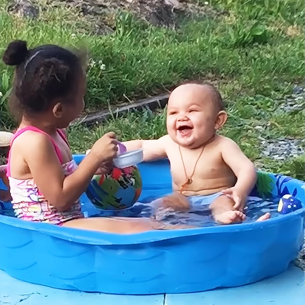 A baby and a young girl are playing in a small inflatable pool, with the baby laughing and the girl holding a toy.
