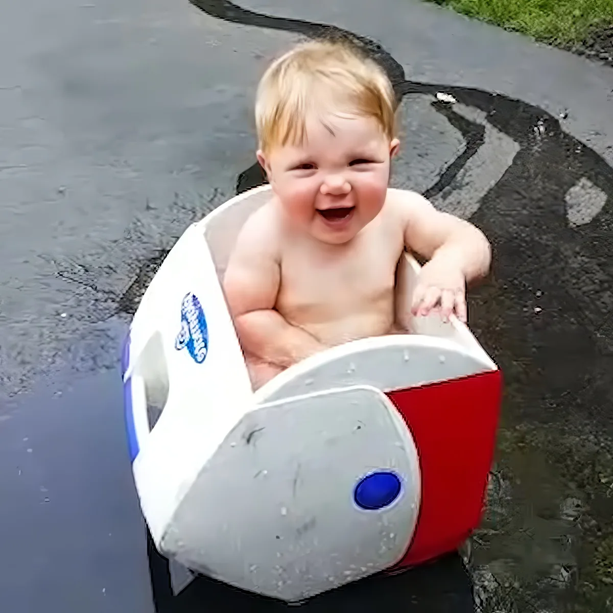 A smiling baby sits in a red and white plastic bucket on a wet pavement.