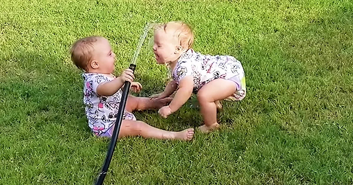 Two babies playfully interact with a garden hose on a grassy lawn.