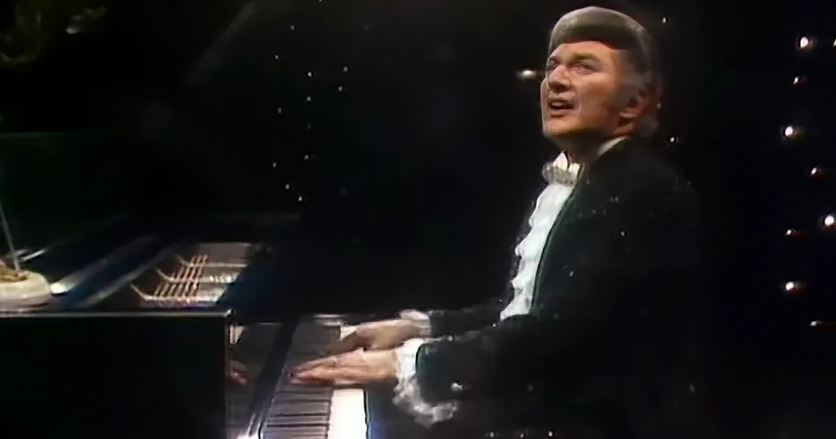 A man in a black suit and hat sings while playing a piano, surrounded by dark background.