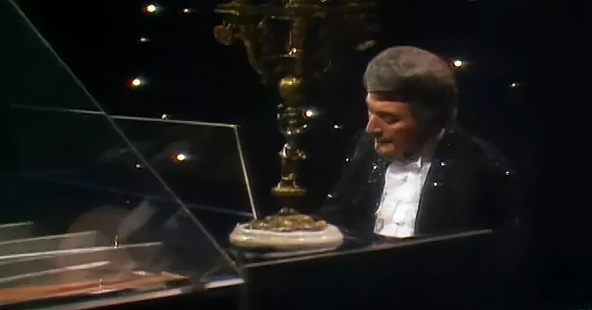 A man in formal attire sits at a grand piano, focused on playing, with a decorative chandelier in the background.