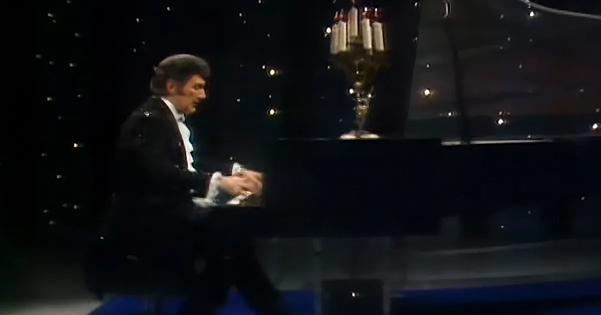 A man in a tuxedo plays a grand piano on a stage with a chandelier.