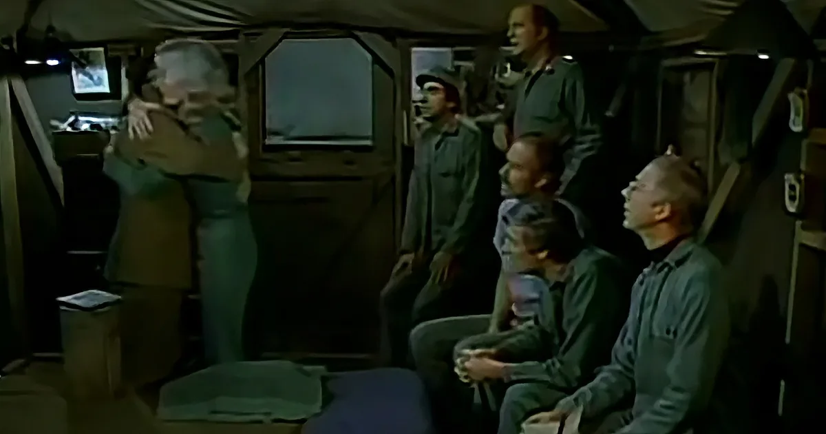 A man in a brown jacket embraces another man in a military setting with others observing.