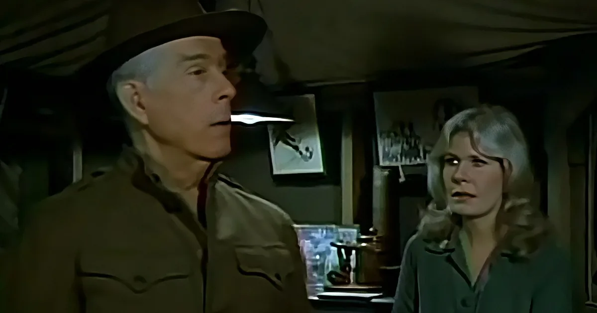 A man in a military uniform stands in a tent, speaking to a woman with blonde hair.