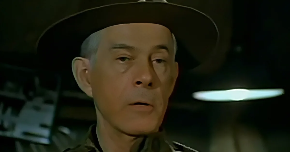 A man wearing a hat and a uniform shirt, looking slightly to the side with a serious expression.