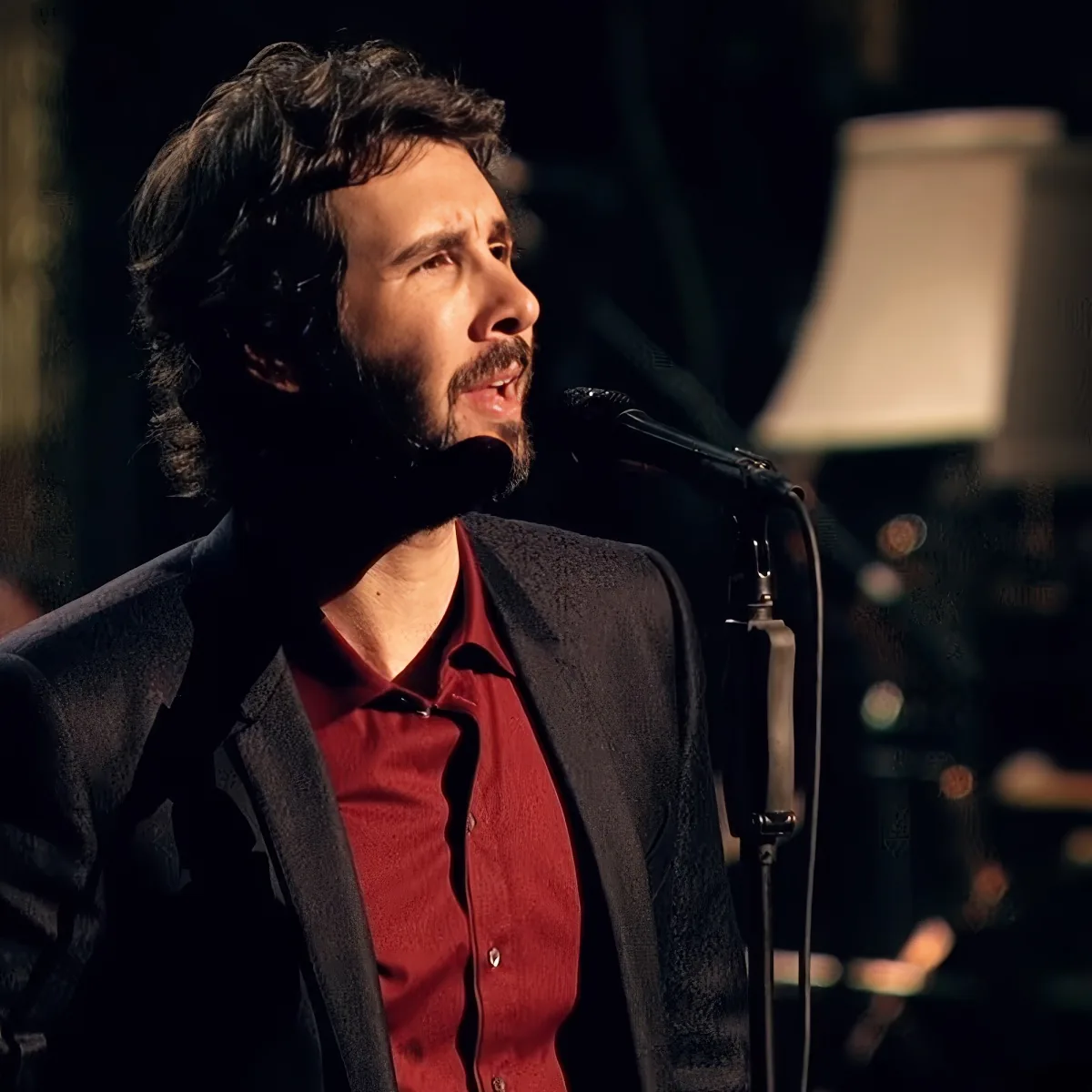 A man with dark hair and a beard sings into a microphone, wearing a red shirt and black jacket.