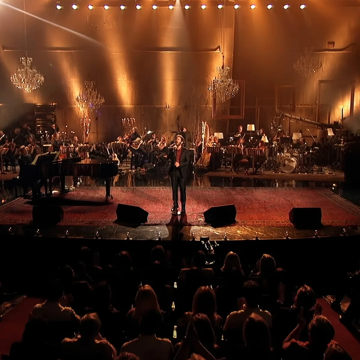A man in a suit sings on stage with a grand piano and orchestra behind him.