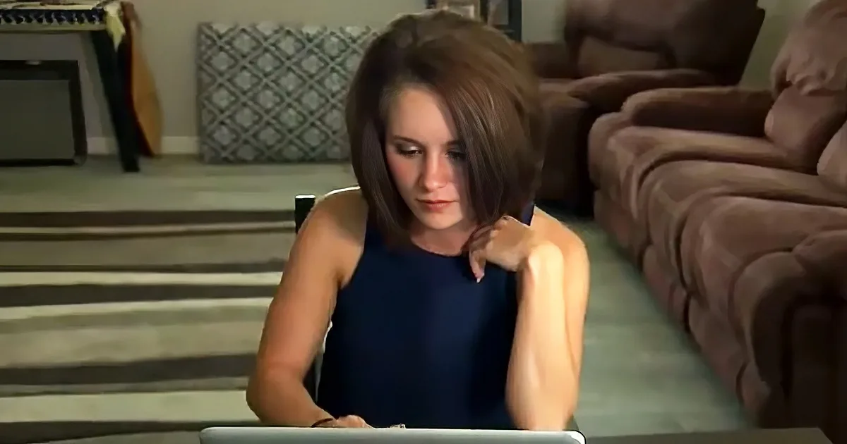 A woman with short brown hair sits in a chair, looking down, wearing a blue sleeveless top.