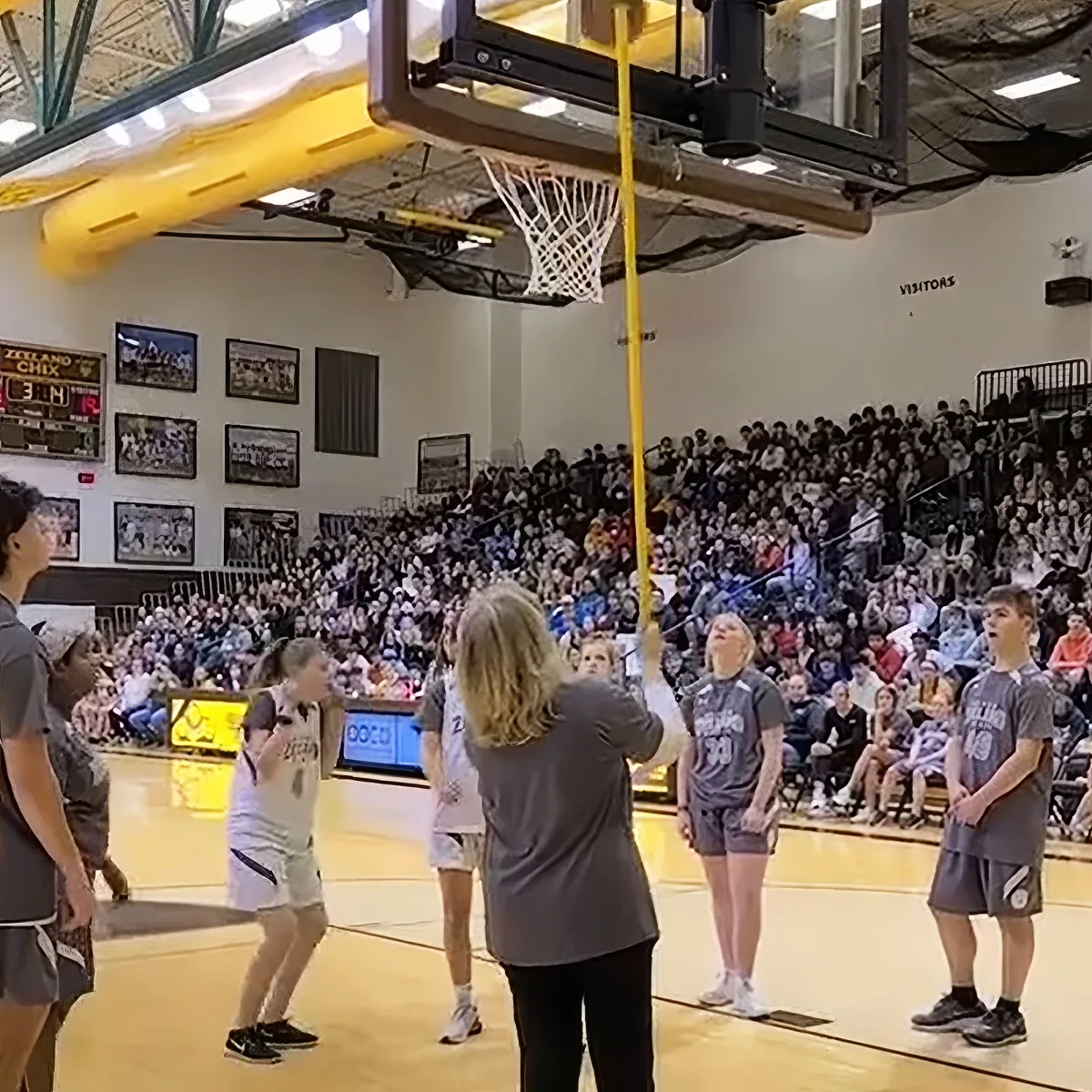 A woman in a gray shirt holds a basketball hoop, with players and spectators in a gymnasium.