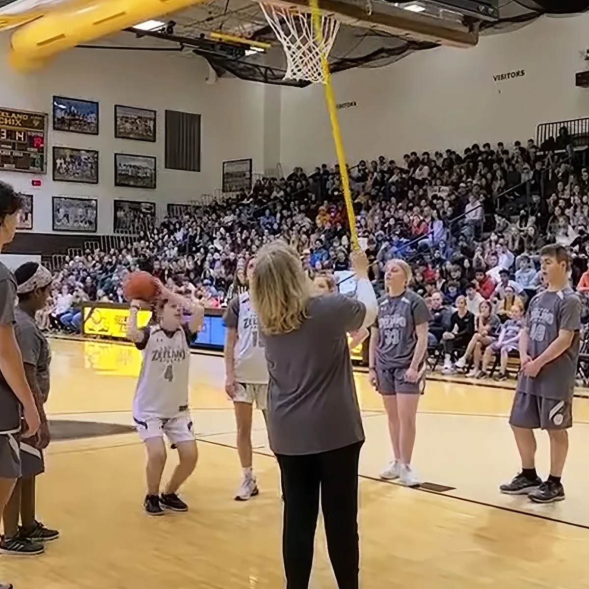 A woman in a gray shirt holds a basketball as a crowd watches in a gymnasium.