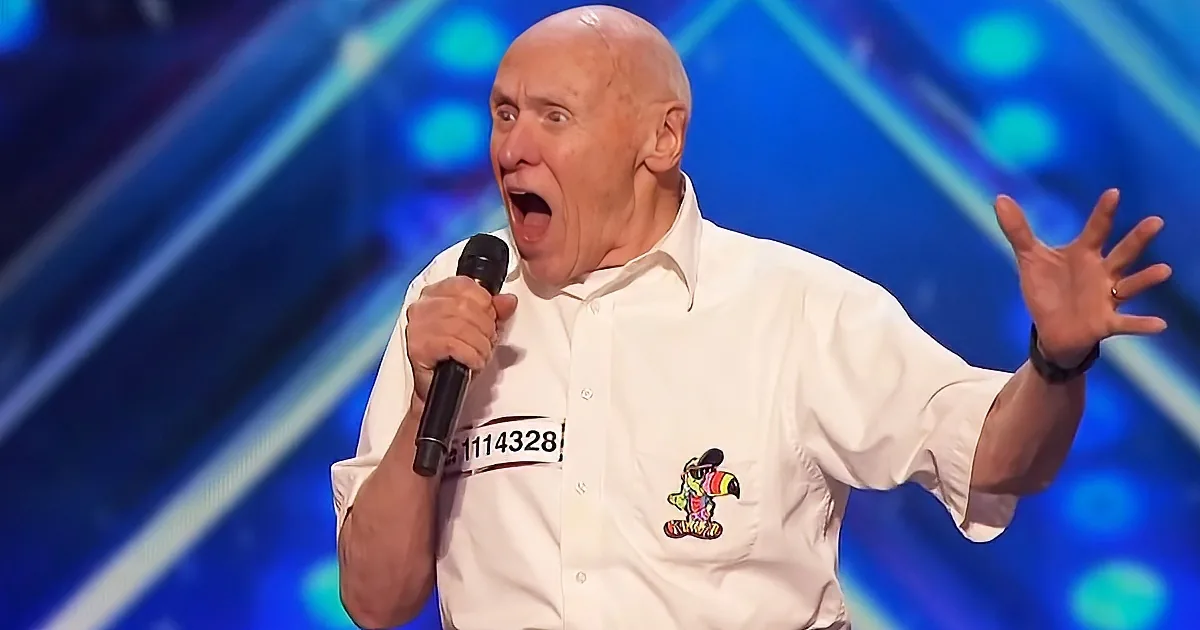 An older man in a white shirt holds a microphone, gesturing with his hands, against a blue stage backdrop.