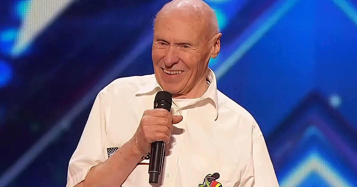 An older man in a white shirt smiles while holding a microphone on a stage with a blue background.