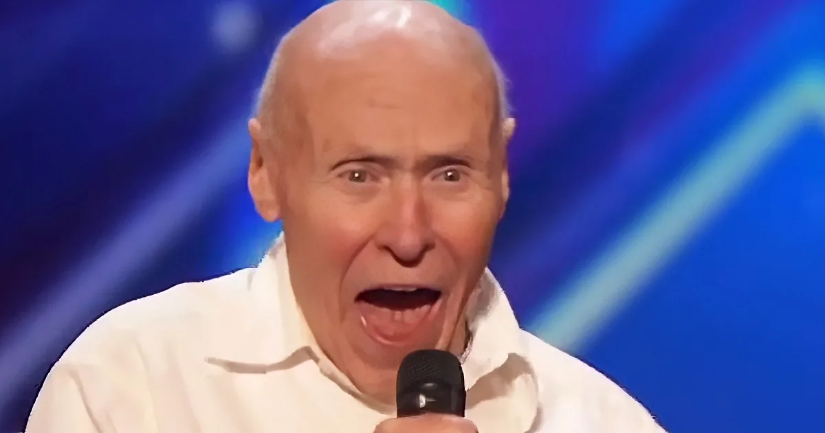 A person with a bald head, wearing a white shirt, is holding a microphone and appears to be speaking or singing.