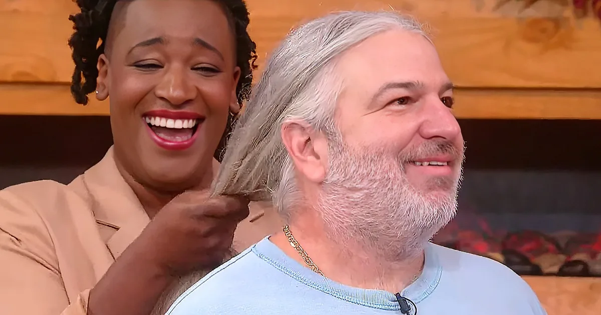 A man with a gray beard and a woman with red lipstick are laughing together, the woman adjusting the man's hair.