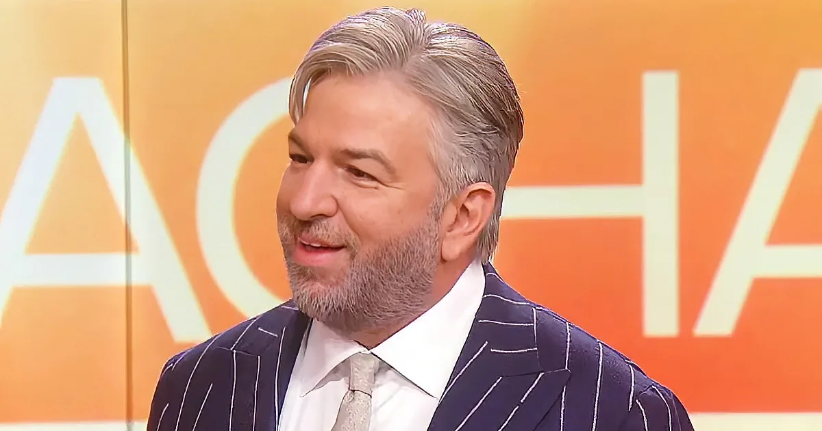 A man with gray hair and a beard, wearing a navy pinstripe suit, is speaking in front of an orange background with large...