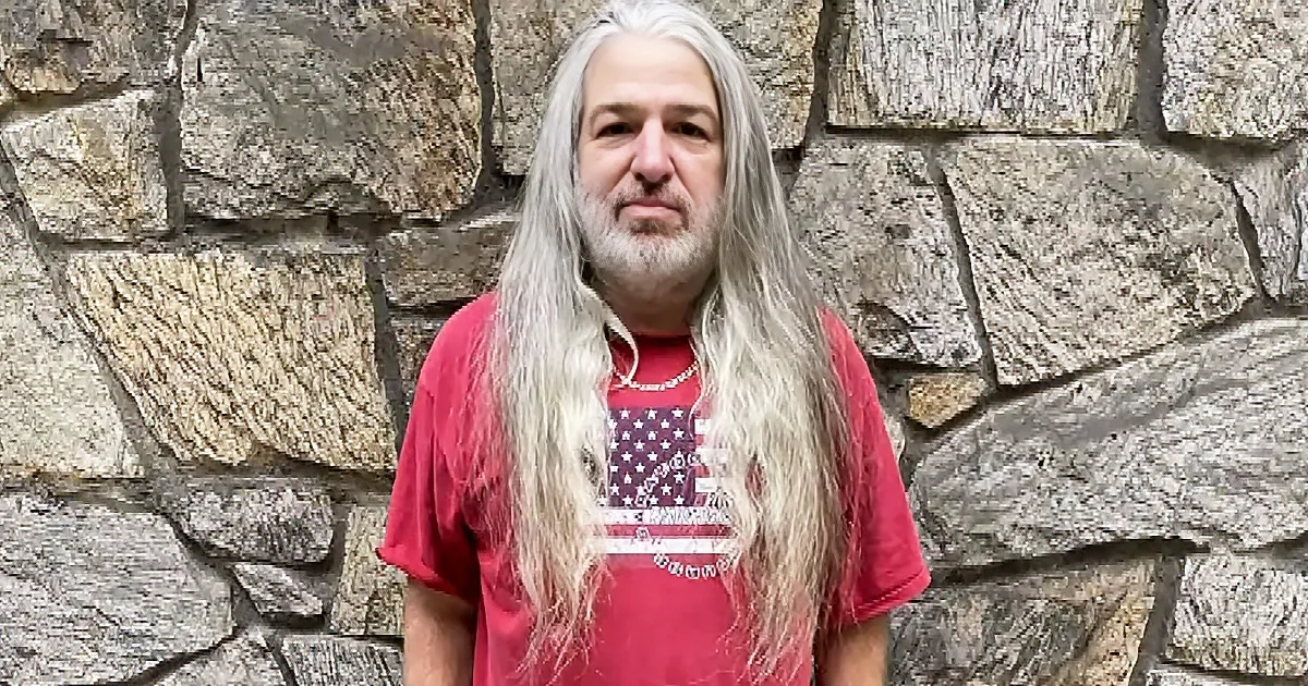 A man with long white hair and a beard stands in front of a stone wall, wearing a red t-shirt with an American flag design.