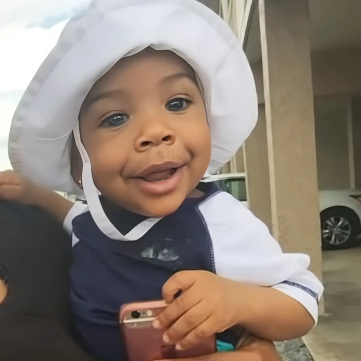 A smiling toddler in a white chef's hat holds a phone, wearing a navy and white outfit.