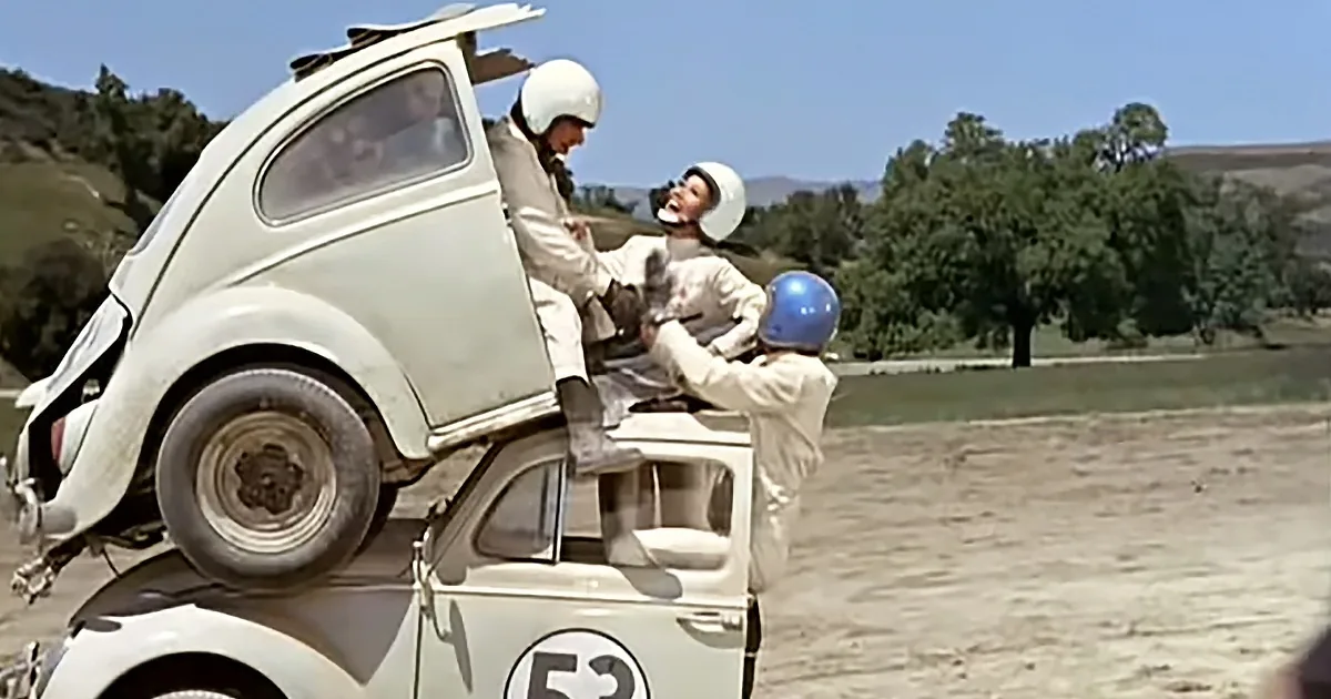 A vintage Volkswagen Beetle with a damaged rear is being worked on by three individuals in racing suits and helmets.