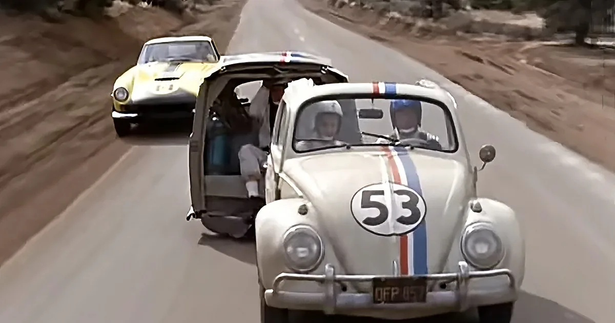 A white Volkswagen Beetle with number 53 races down a road, followed by a yellow sports car.