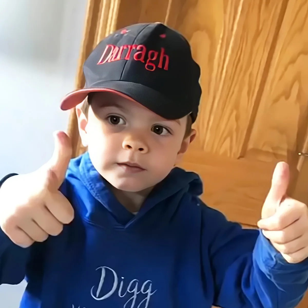 A child wearing a blue hoodie and a black cap with "Durragh" on it gives two thumbs up.