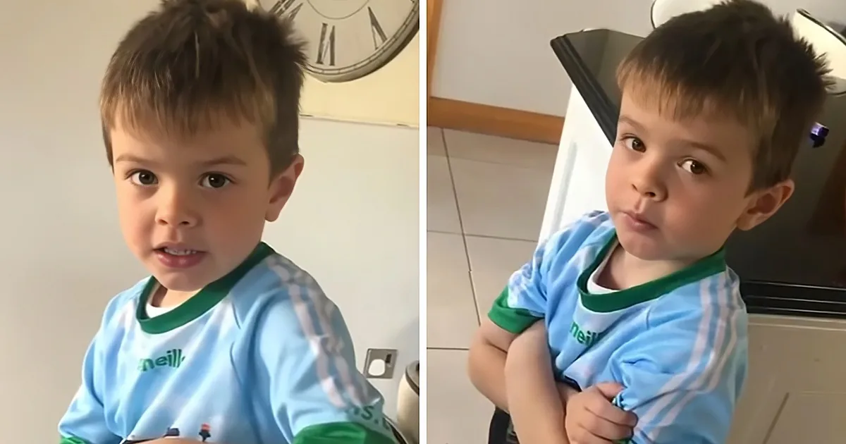 A young boy in a blue and green shirt stands indoors, looking slightly to the side.