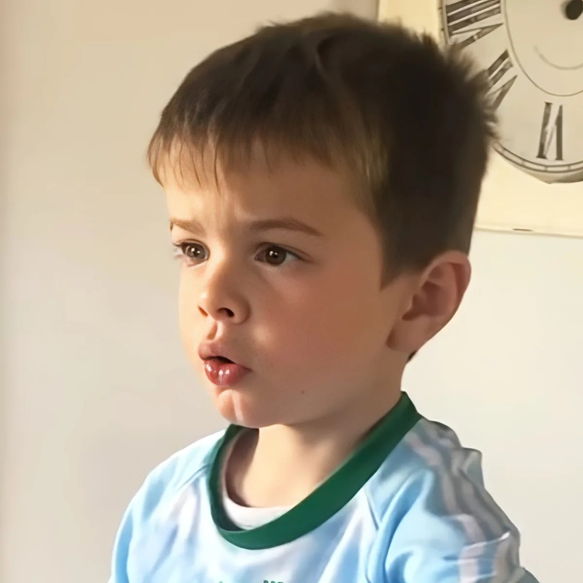 A young boy with short brown hair, wearing a light blue shirt with a green collar, looks to the side with a curious...