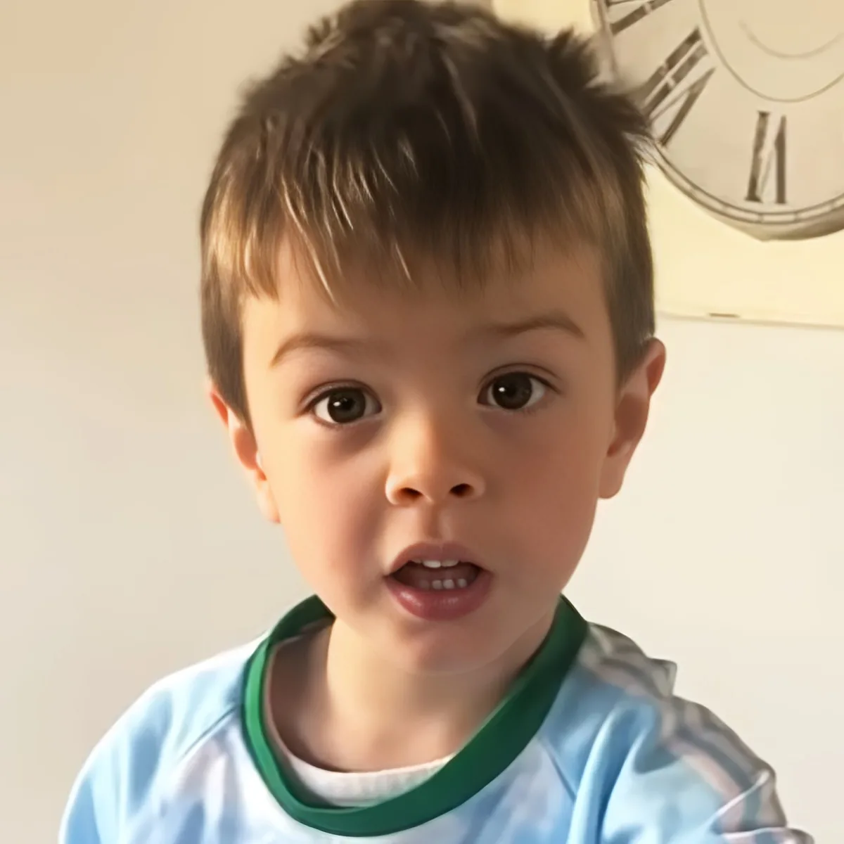 A young child with short brown hair wearing a light blue shirt with green trim is looking directly at the camera.