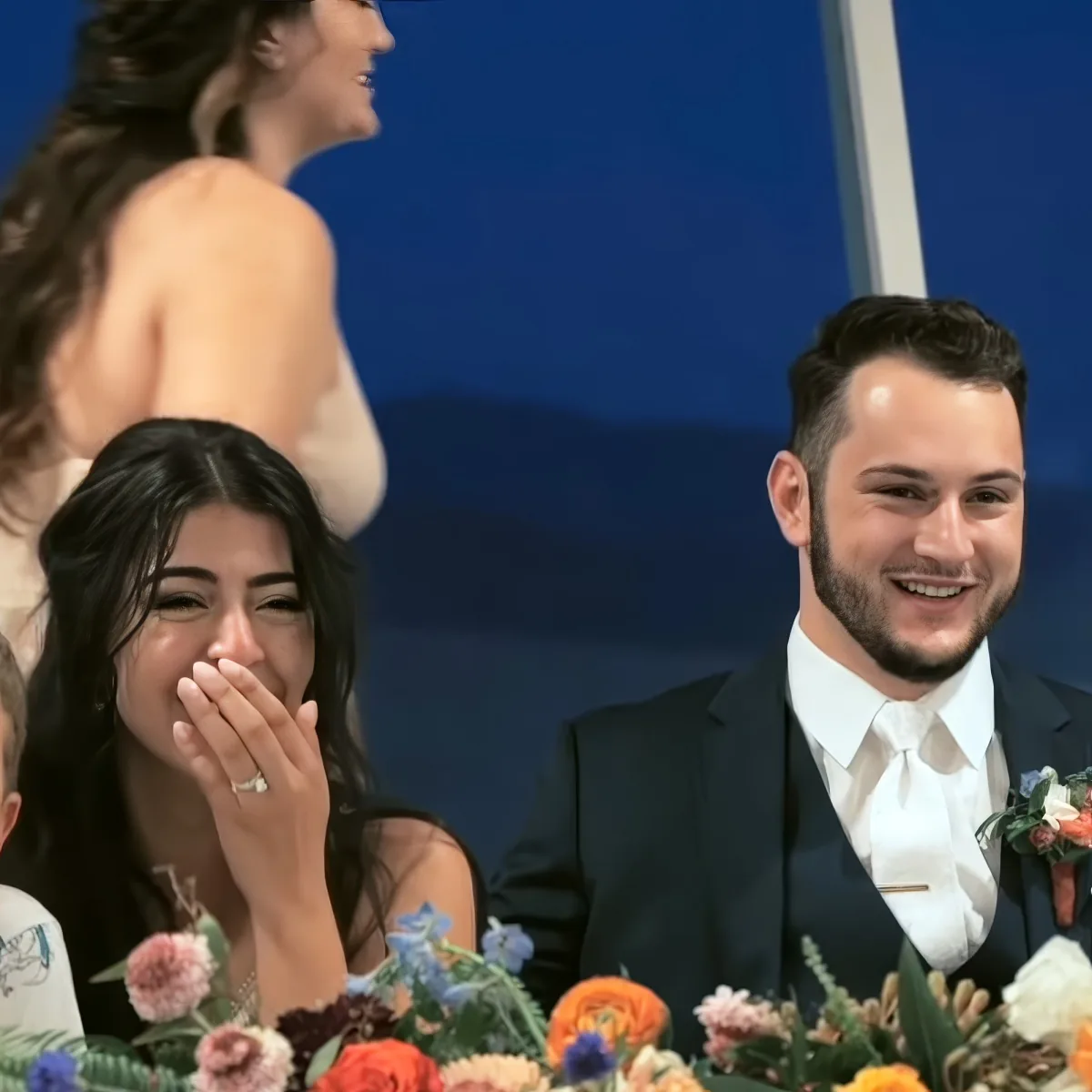 A bride laughs while a groom smiles at a wedding reception, surrounded by floral decorations.