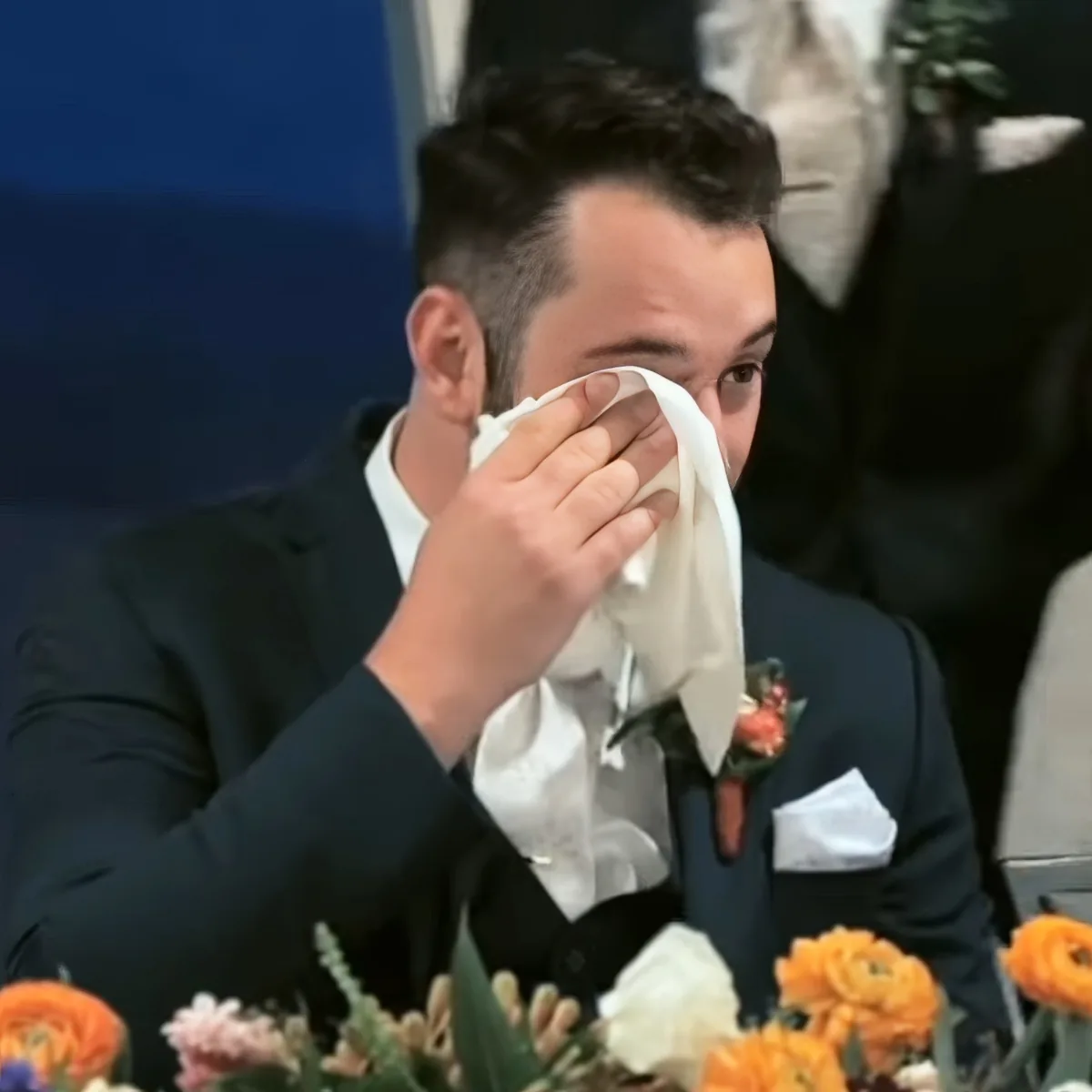A man in a suit wipes his face with a tissue, surrounded by flowers and a formal setting.