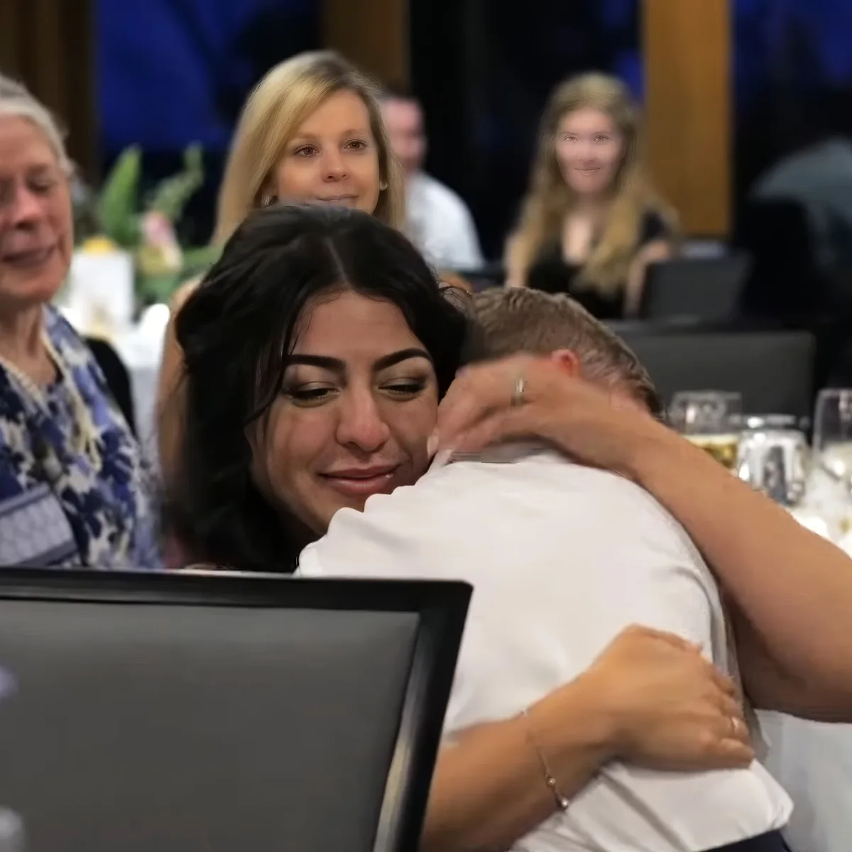 A woman is hugging a man at a formal event, surrounded by other attendees.