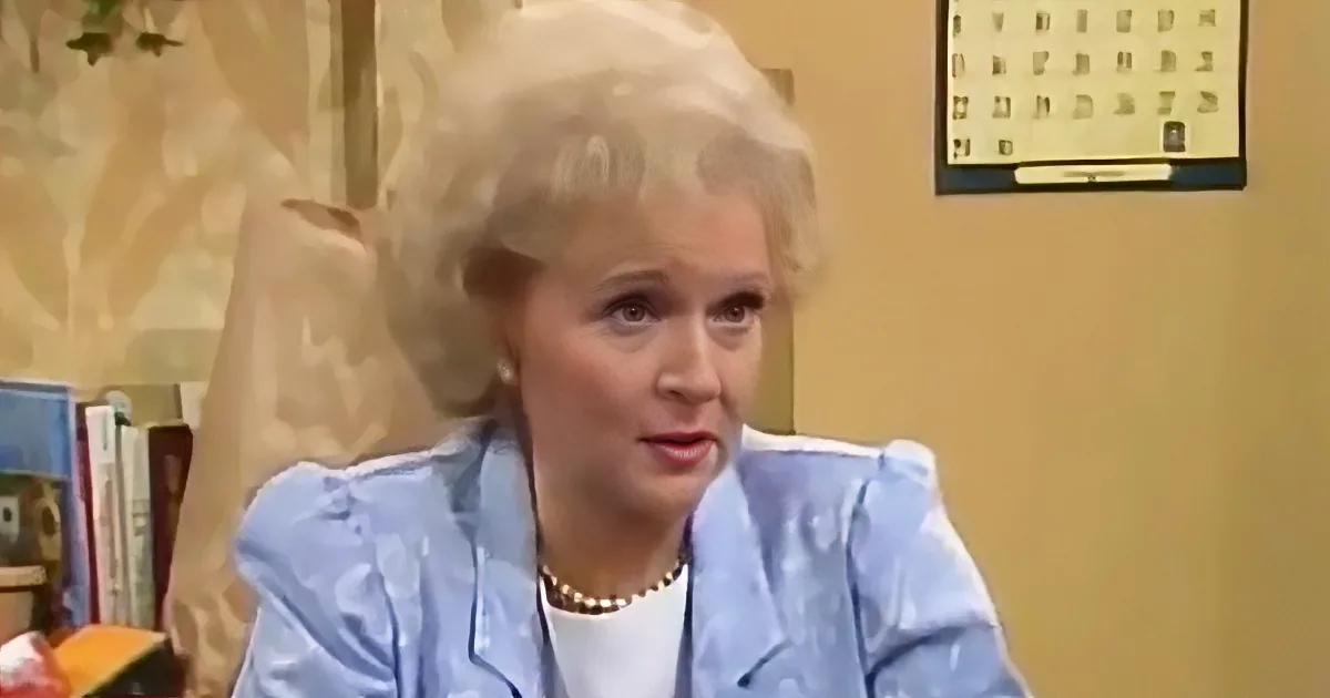 A woman with white hair, wearing a blue floral jacket and pearl necklace, sits in a room with a calendar on the wall.