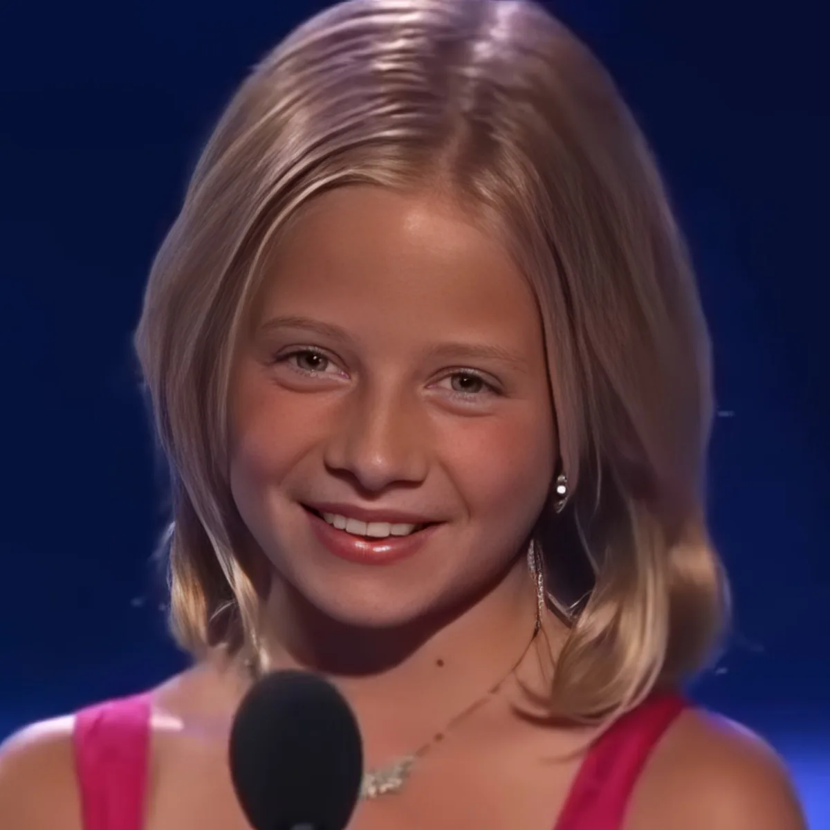 A young girl with blonde hair smiles while holding a microphone, wearing a pink top and a necklace.