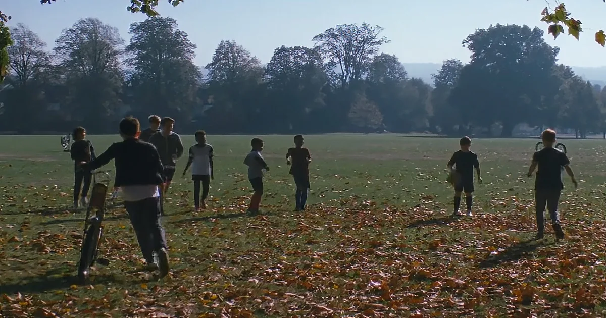 Children play in a park with fallen leaves, a bicycle nearby, and trees in the background.