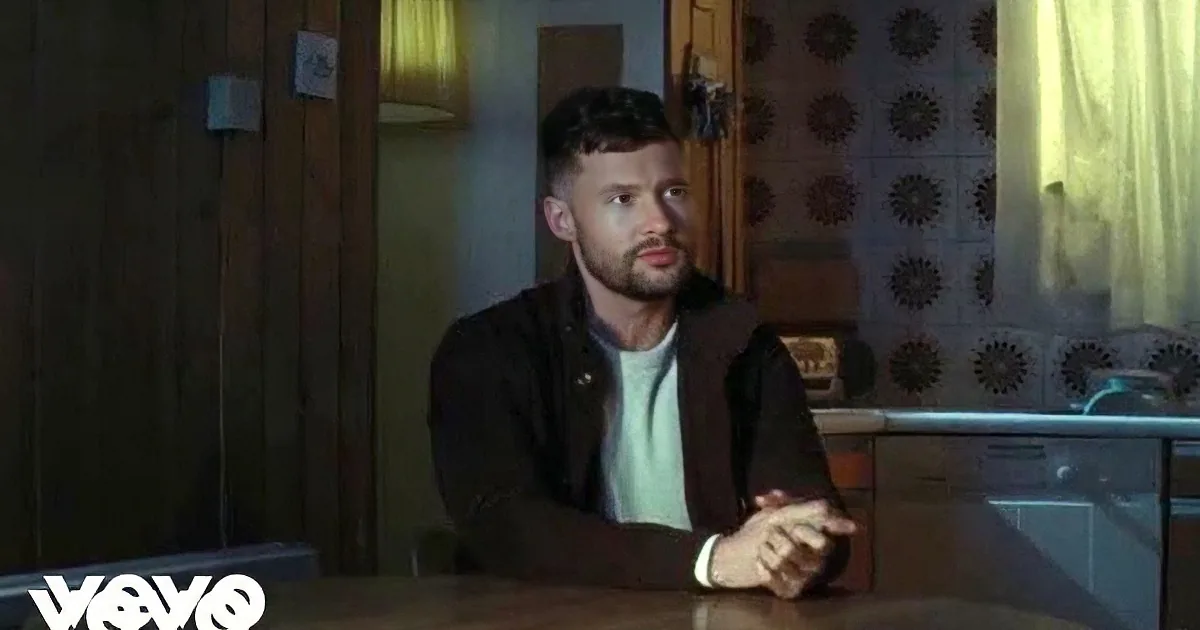 A man with a beard sits at a wooden table in a dimly lit kitchen, hands clasped.