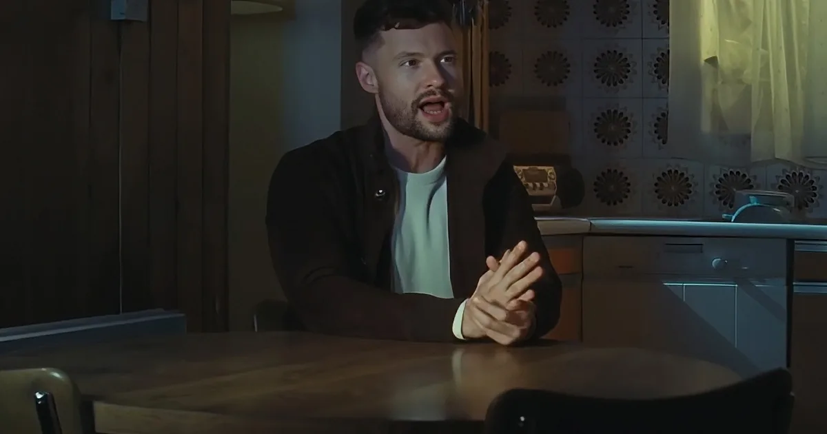 A man sits at a table in a dimly lit kitchen, clasping his hands and appearing surprised or shocked.