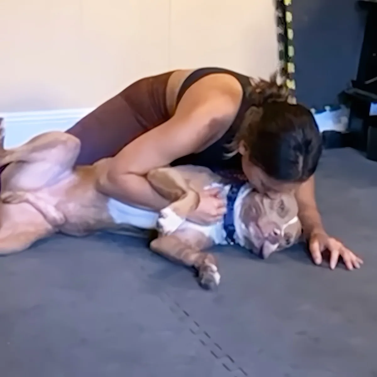 A woman in a brown tank top is playfully wrestling with a dog on a gray mat.