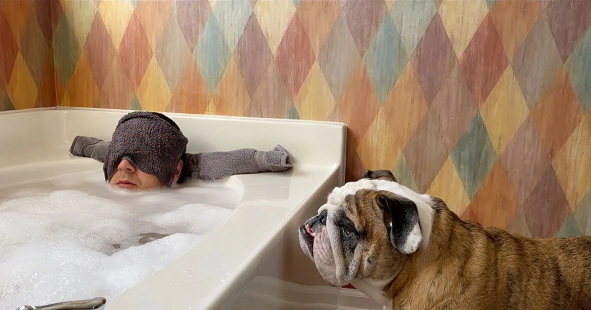 A man in a bathtub with a dog looking at him, both surrounded by colorful diamond-patterned tiles.