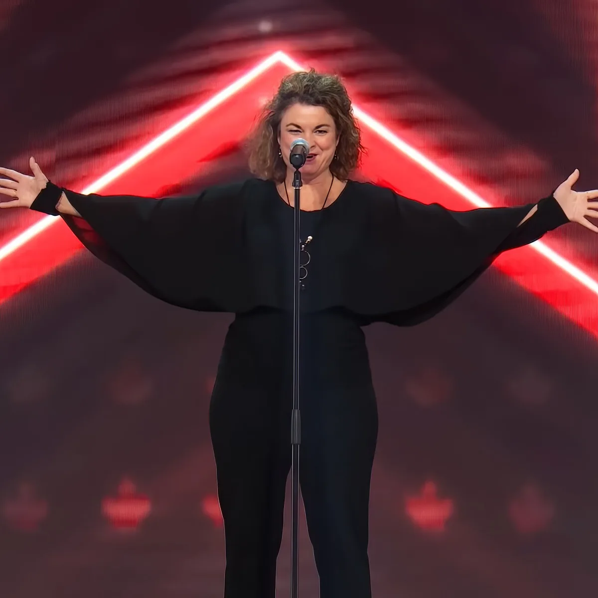 A woman in a black outfit stands on stage with arms outstretched, singing into a microphone.