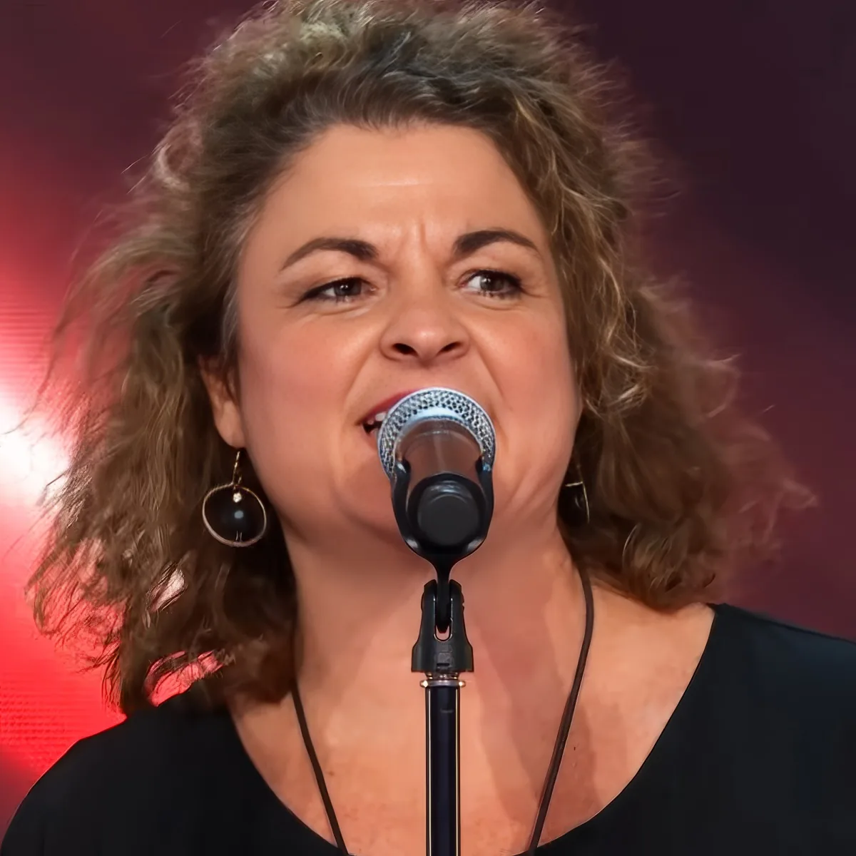 A woman with curly hair and earrings sings into a microphone against a red background.