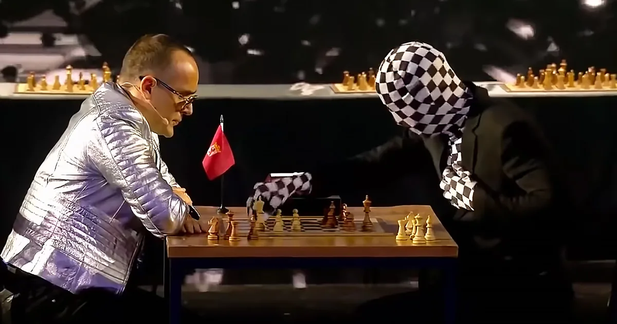 A man in a shiny jacket plays chess with a person wearing a checkered headscarf, both focused on the game.