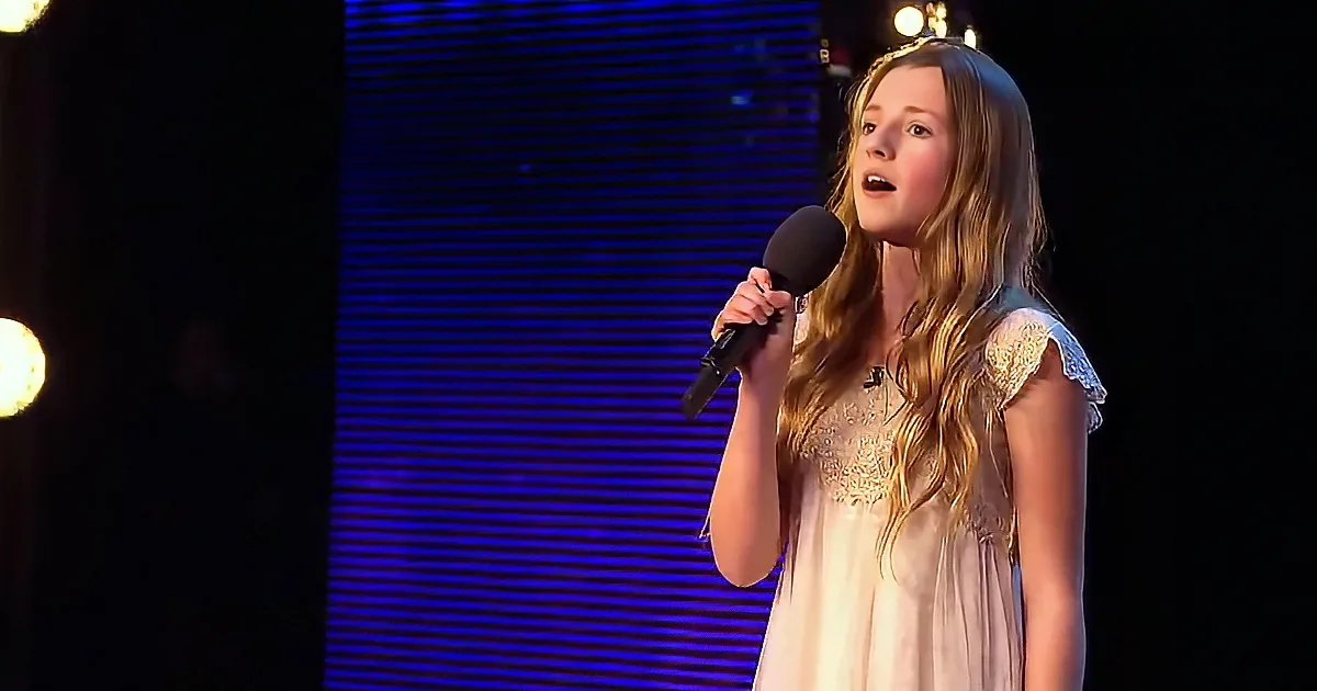 A young girl in a white dress sings into a microphone on stage with blue lighting.