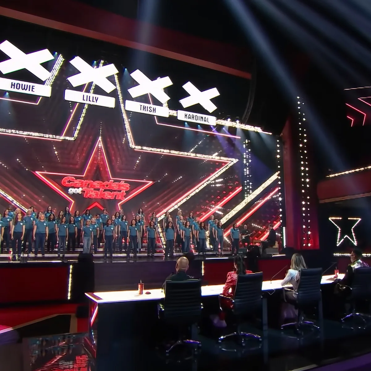 A choir in blue shirts performs on a stage with judges' tables in front.